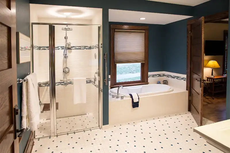 A bright, modern bathroom with a glass-enclosed shower, a white soaking tub next to a wood-trimmed window, and teal walls with decorative tile accents.